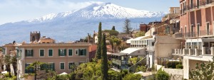 Panoramic view of historic Sicilian town of Taormina, in the background with white peak of volcano Etna with smoke on hot spring day